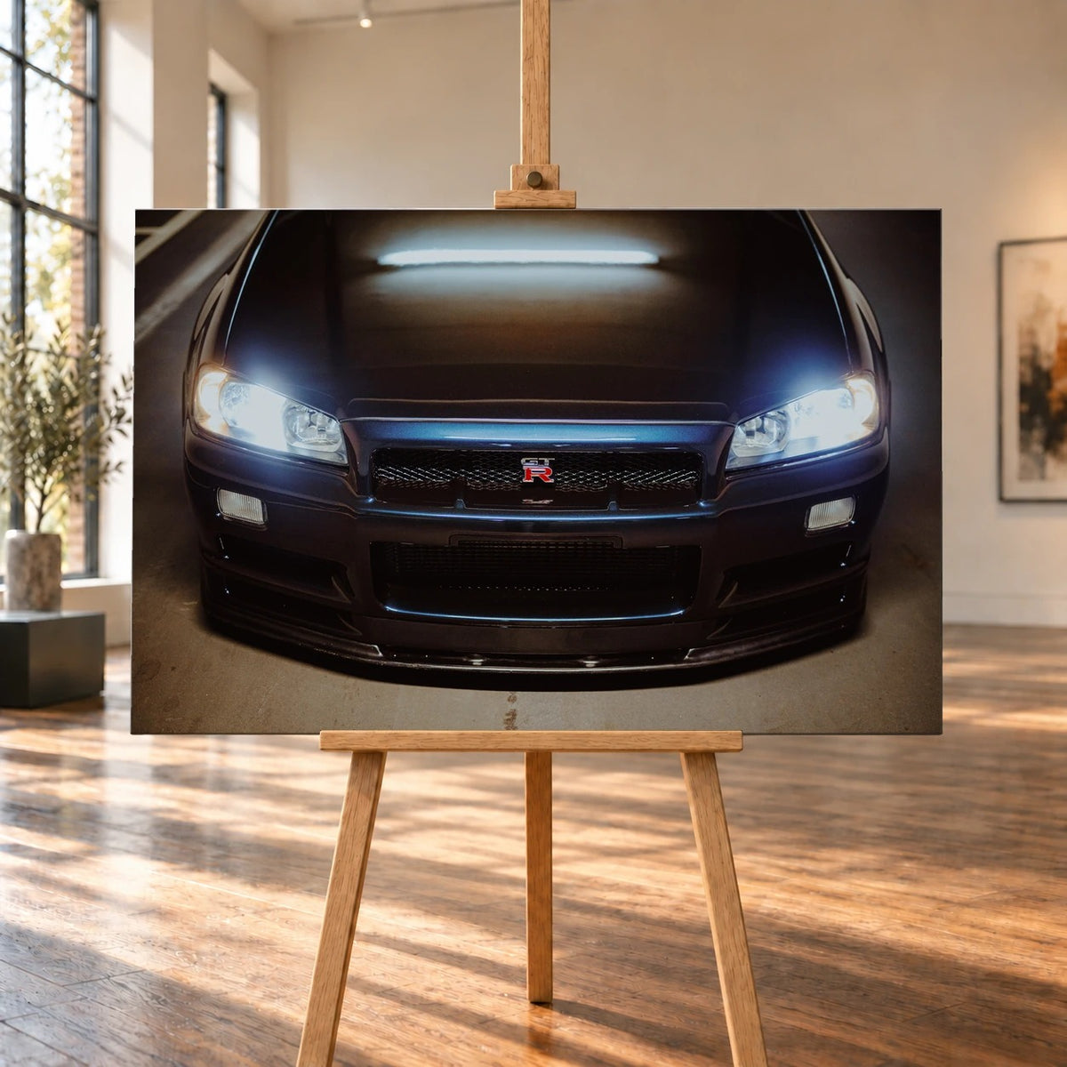 Raumvisualisierung mit einem querformatigen Leinwandbild auf einer Holzstaffelei in einem hellen Raum mit großen Fenstern und Holzboden. Die frontal dargestellte dunkelblaue Fahrzeugfront des Nissan Skyline GT-R R34 füllt das Bild; beide Scheinwerfer leuchten.