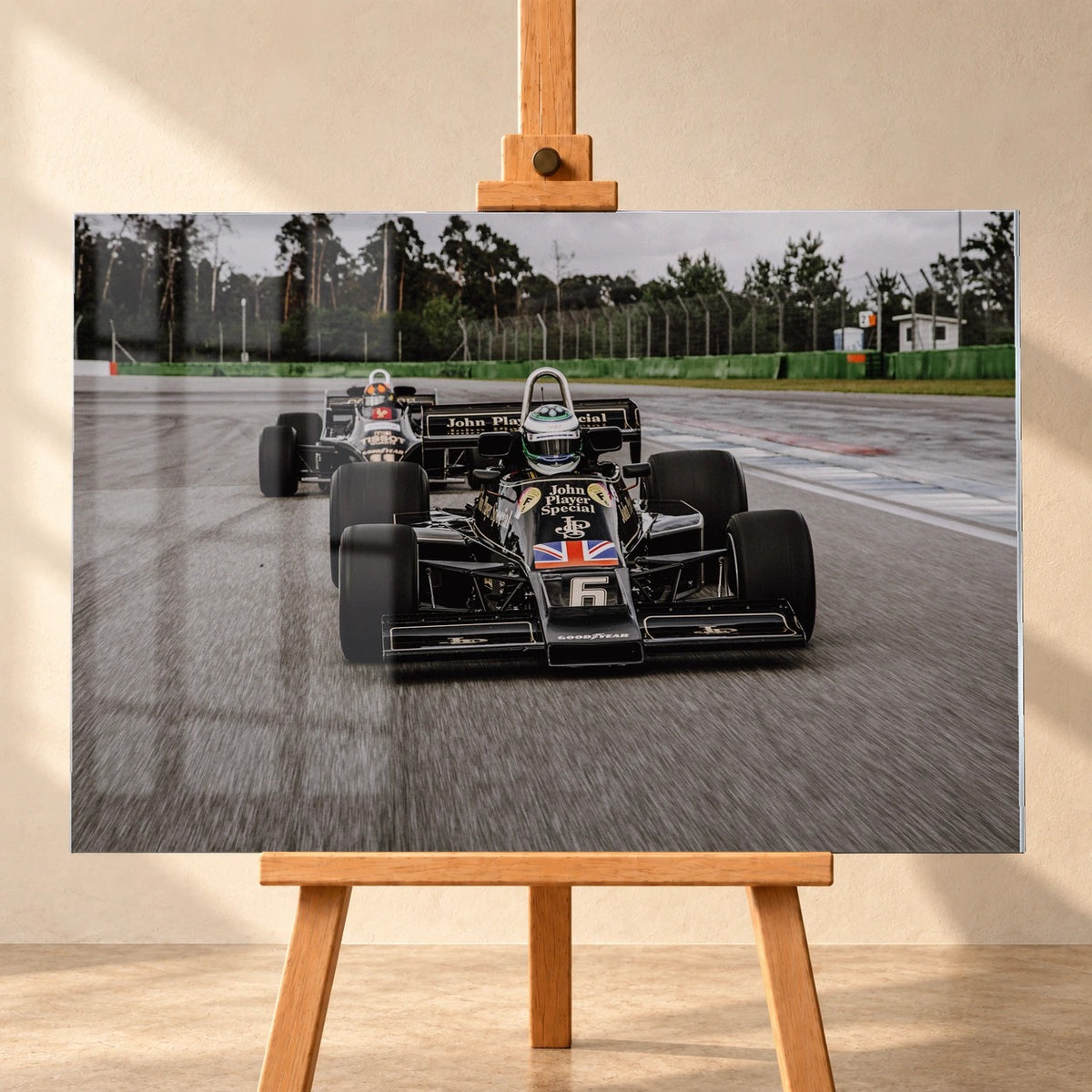 Visualisierung eines Acrylglasbildes im Querformat, frontal auf einer Holzstaffelei vor heller Wand. Über dem Motiv mit Lotus 77 und Lotus 88 auf der Rennstrecke sind links Spiegelungen dargestellt; der schwarz-goldene Wagen mit Startnummer 6 dominiert den Vordergrund.