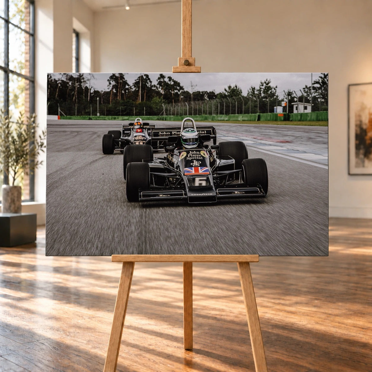 Raumvisualisierung mit einem querformatigen Leinwandbild auf einer Holzstaffelei zwischen großen Fenstern und Holzboden. Auf dem Bild sind Lotus 77 und Lotus 88 in schwarz-goldener Lackierung auf der Rennstrecke zu sehen; der Wagen mit Startnummer 6 steht im Vordergrund.