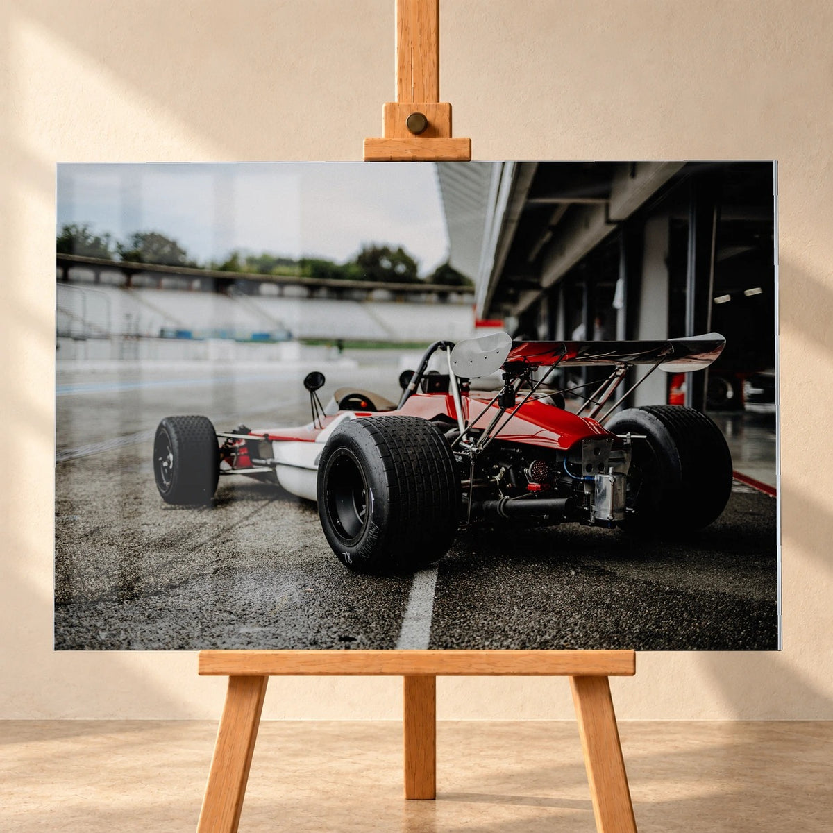 Frontalansicht eines visualisierten Acrylglas-Wandbilds auf einer Holzstaffelei vor heller Wand. Das Motiv zeigt den rot-weißen Lotus 69 Formel 2 von schräg hinten auf nassem Asphalt, mit breiten Hinterreifen und hohem Heckflügel.