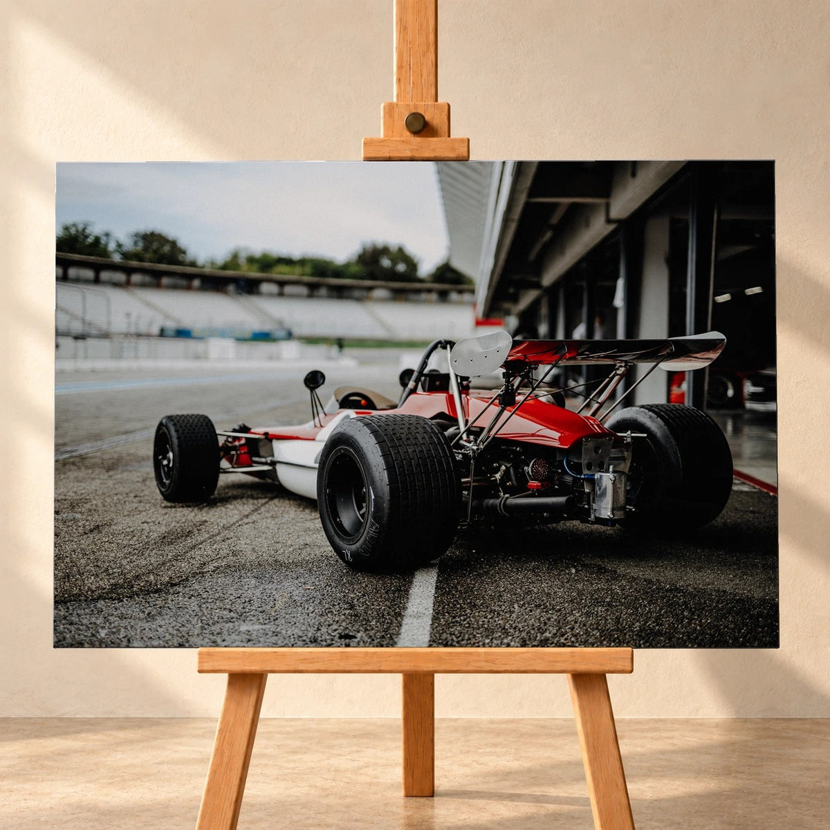 Visualisierung eines querformatigen Leinwandbilds auf einer Holzstaffelei vor heller Wand. Das Motiv zeigt den rot-weißen Lotus 69 Formel 2 von schräg hinten mit breiten Hinterreifen und hohem Heckflügel in der Boxengasse.