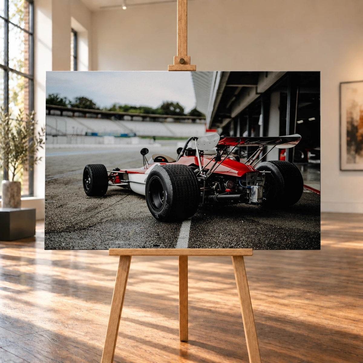 Raumvisualisierung mit einem Leinwandbild auf einer Holzstaffelei in einem hellen Raum mit großen Fenstern und Holzboden. Auf dem querformatigen Bild ist der rot-weiße Lotus 69 Formel 2 von schräg hinten in der Boxengasse zu sehen.
