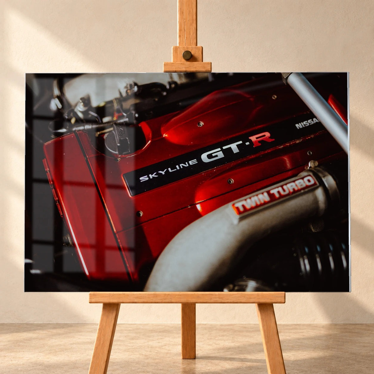 Frontal dargestelltes Acrylglasbild auf einer Holzstaffelei vor heller Wand, als Produktvisualisierung mit sichtbarer Fensterspiegelung. Das Motiv zeigt die rote Motorabdeckung eines Nissan Skyline GT-R R34 und ein silberfarbenes Rohr mit „TWIN TURBO“-Aufschrift.