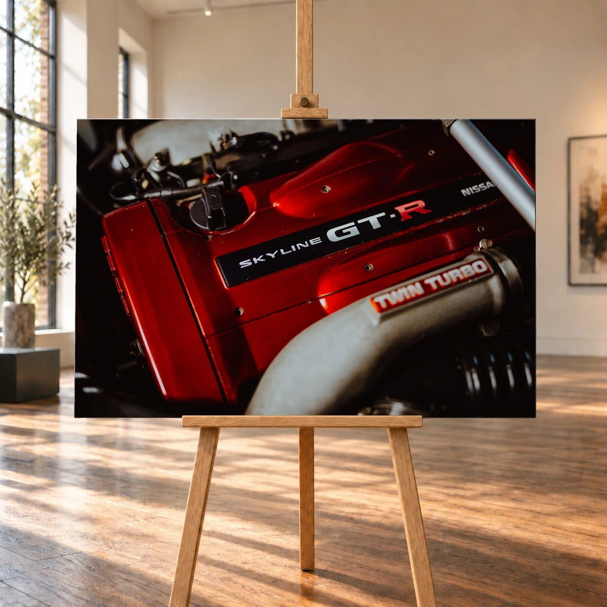 Raumvisualisierung mit einem querformatigen Leinwandbild auf einer Holzstaffelei in einem hellen Raum mit hohen Fenstern und Holzboden. Das Bild zeigt eine Nahaufnahme der roten Motorabdeckung eines Nissan Skyline GT-R R34 mit „SKYLINE GT-R“-Schriftzug.