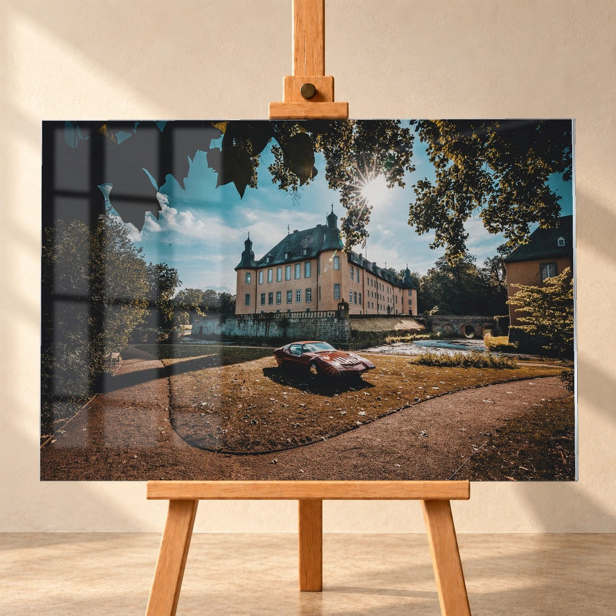Frontal visualisiertes Acrylglasbild auf einer Holzstaffelei vor heller Wand, mit Fensterspiegelung auf der linken Bildhälfte. Das Motiv zeigt den roten Pontiac Number One vor einem Schloss unter sonnenbeschienenen Baumkronen.