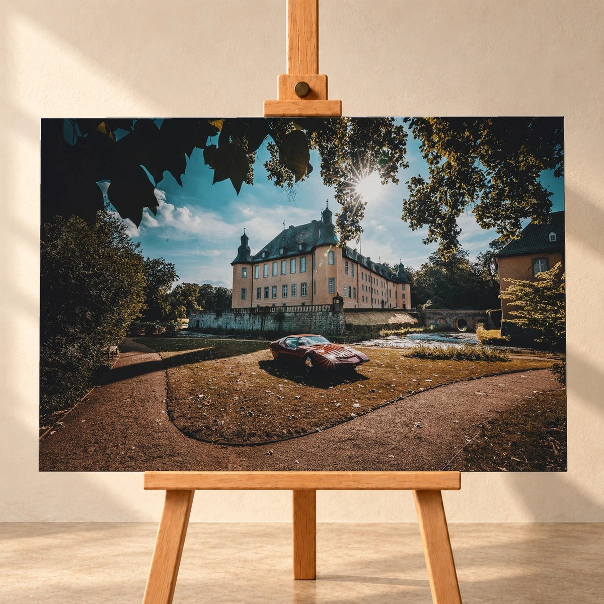 Visualisierung eines querformatigen Leinwandbilds auf einer Holzstaffelei vor heller Wand. Das Motiv zeigt den roten Pontiac Number One auf einer Rasenfläche vor einem Schloss, mit Sonnenlicht zwischen den Baumblättern.