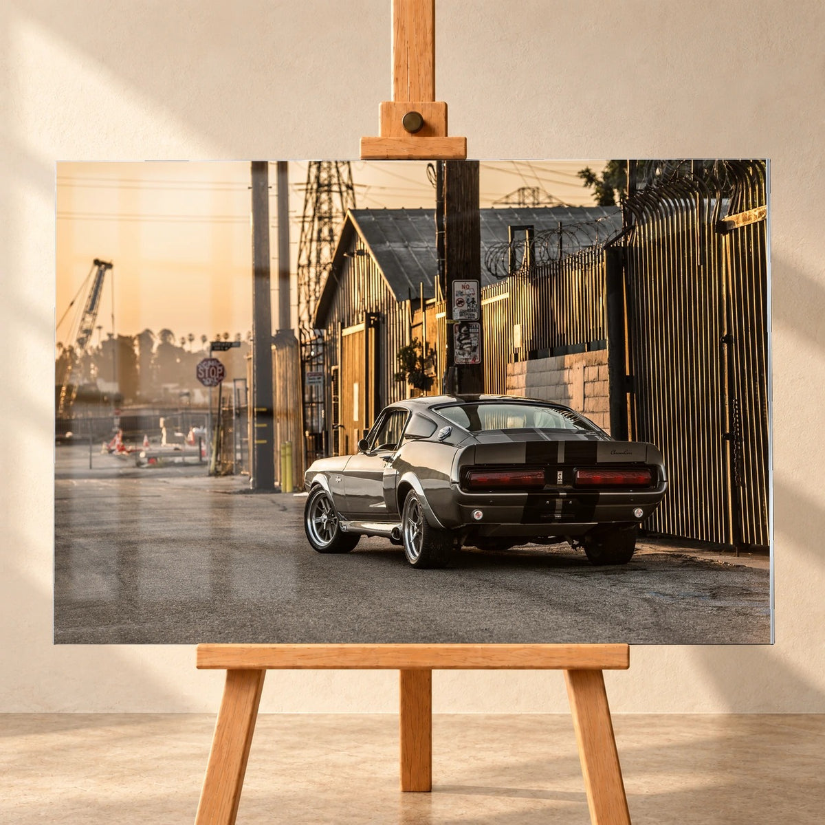 Der graue Ford Mustang Shelby GT500 „Eleanor“ mit schwarzen Streifen, schräg von hinten an einem Metallzaun im goldenen Sonnenlicht. Das Motiv ist als querformatiges Acrylglasbild frontal auf einer Holzstaffelei vor heller Wand visualisiert.