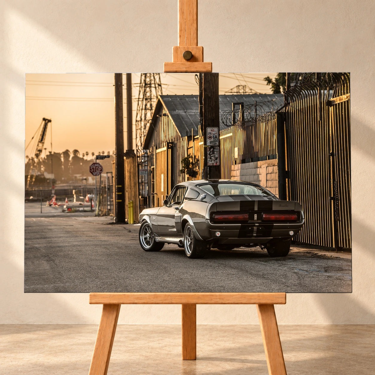 Frontale Visualisierung eines Leinwandbilds im Querformat auf einer Holzstaffelei vor heller Wand. Das Motiv zeigt den grauen Ford Mustang Shelby GT500 „Eleanor“ mit schwarzen Streifen schräg von hinten neben einem Industriezaun im warmen Sonnenlicht.
