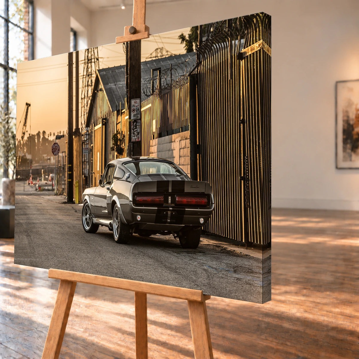 Schrägansicht eines Leinwandbilds als Produktvisualisierung auf einer Holzstaffelei. Das Motiv setzt sich über die sichtbare rechte Seitenkante fort und zeigt den grauen Ford Mustang Shelby GT500 „Eleanor“ von schräg hinten neben einem hohen Metallzaun.