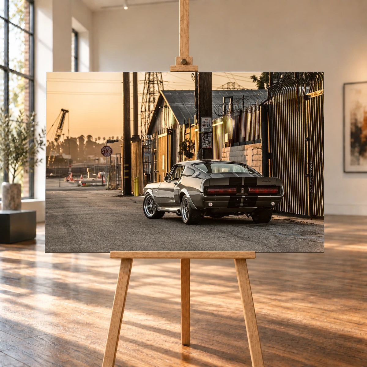 Raumvisualisierung mit einem Leinwandbild auf einer Holzstaffelei in einem hellen Raum mit großen Fenstern und Holzboden. Auf dem querformatigen Bild steht der graue Ford Mustang Shelby GT500 „Eleanor“ in Heckdreiviertelansicht an einem sonnenbeschienenen Industriezaun.
