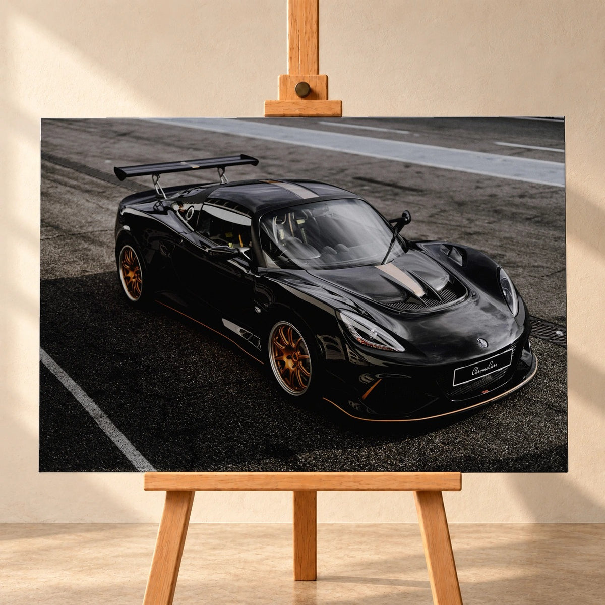 Visualisierung eines Leinwandbilds im Querformat auf einer Holzstaffelei vor heller Wand. Das Motiv zeigt den schwarzen Lotus Exige 430 Cup Type 79 schräg von vorn und oben, mit goldfarbenen Felgen und großem Heckflügel auf Asphalt.