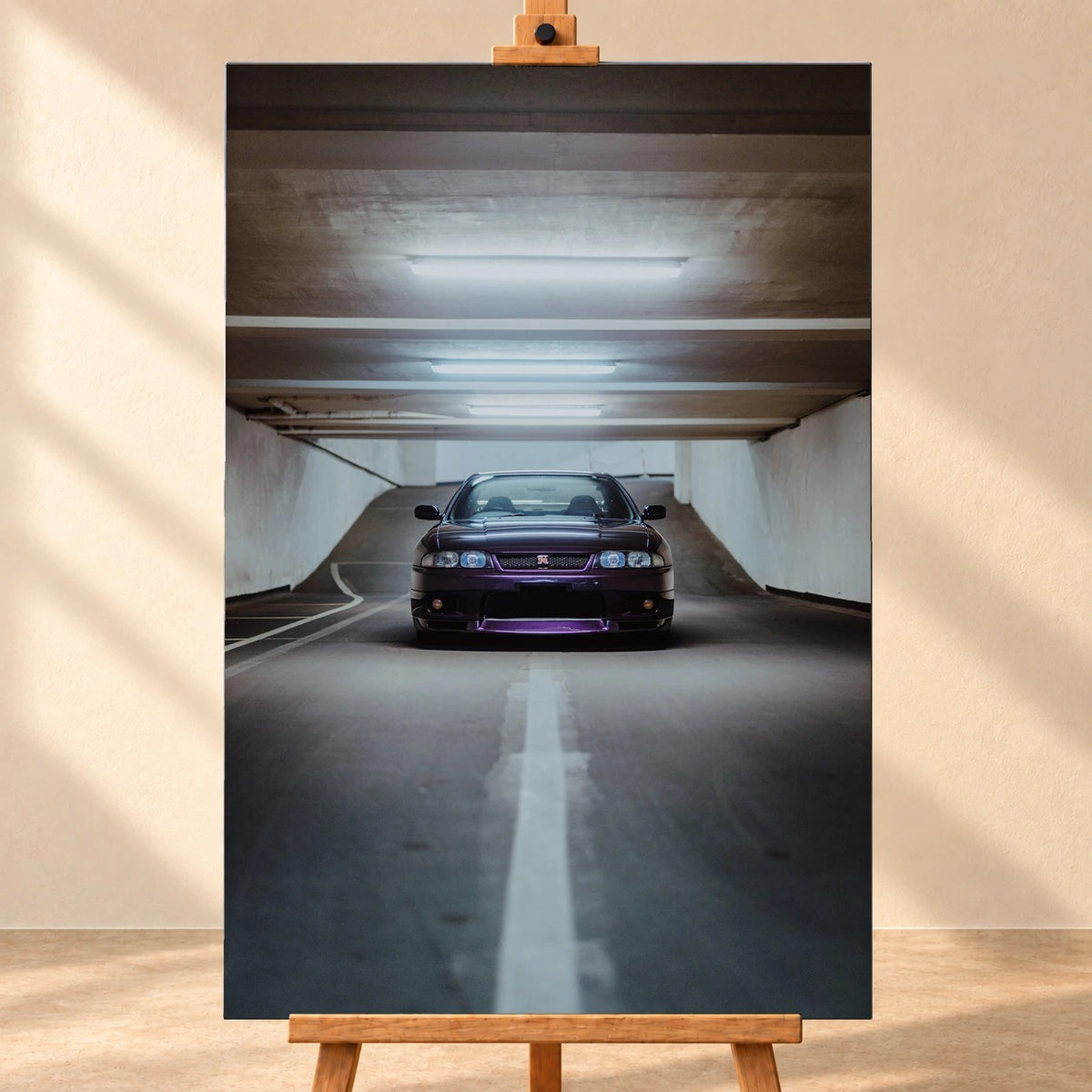 Visualisierung eines hochformatigen Leinwandbilds, frontal auf einer Holzstaffelei vor heller Wand. Das Motiv zeigt einen dunkelvioletten Nissan Skyline GT-R R33 von vorn in einer Tiefgarage unter hellen Deckenleuchten.