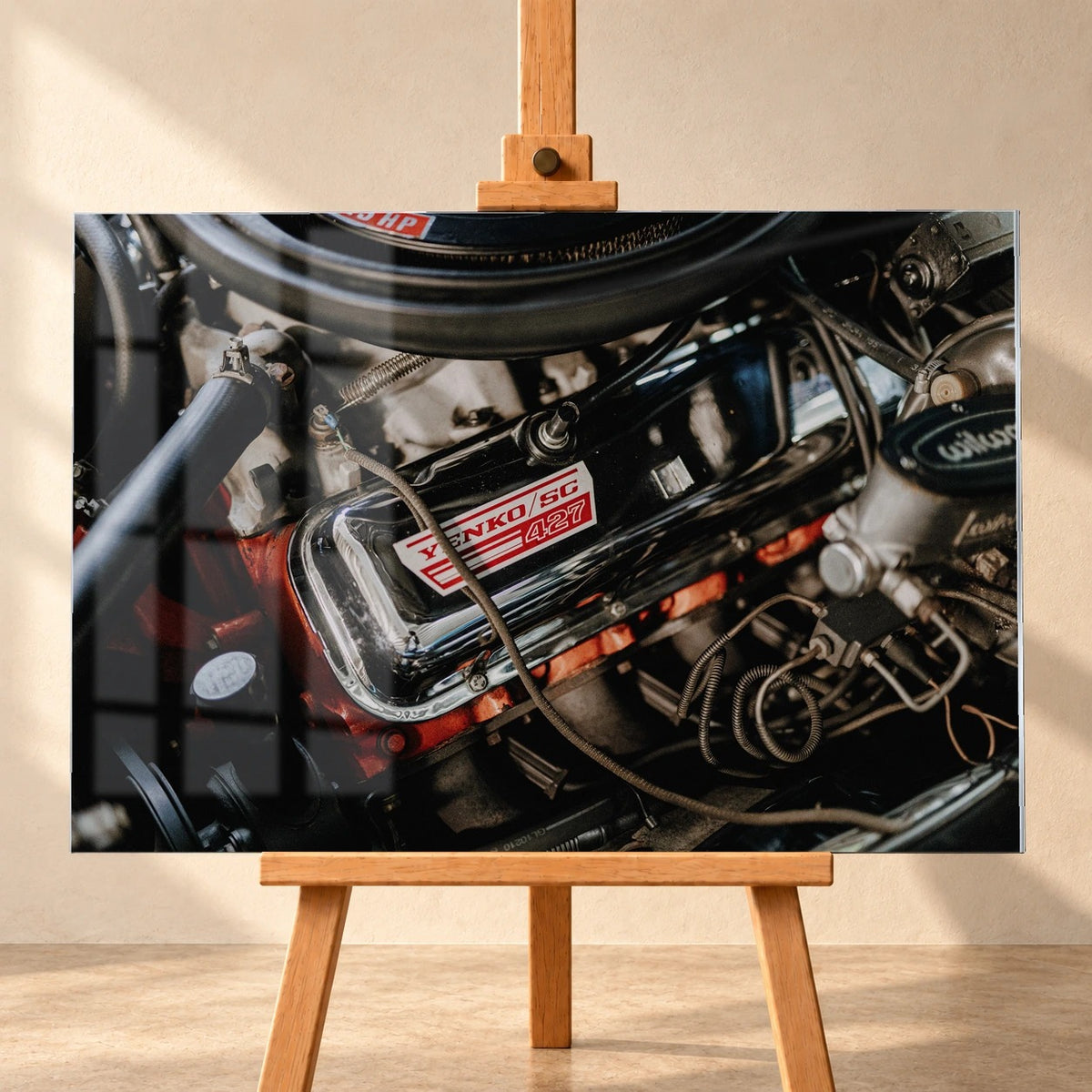 Frontalansicht eines Acrylglasbilds als Visualisierung auf einer Holzstaffelei vor heller Wand. Das Motiv zeigt den Motorraum des Chevrolet Camaro Yenko mit „YENKO/SC 427“ auf dem Ventildeckel; links liegt eine Fensterspiegelung über der Bildfläche.