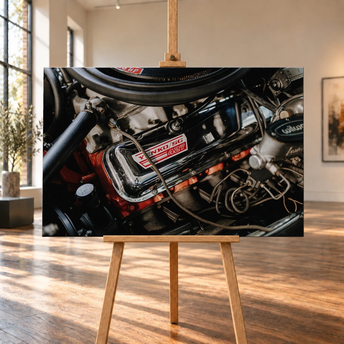 Raumvisualisierung eines Leinwandbilds auf einer Holzstaffelei zwischen hohen Fenstern und Holzboden. Das Motorraummotiv des Chevrolet Camaro Yenko zeigt einen verchromten Ventildeckel mit „YENKO/SC 427“ über dem roten Motorblock.