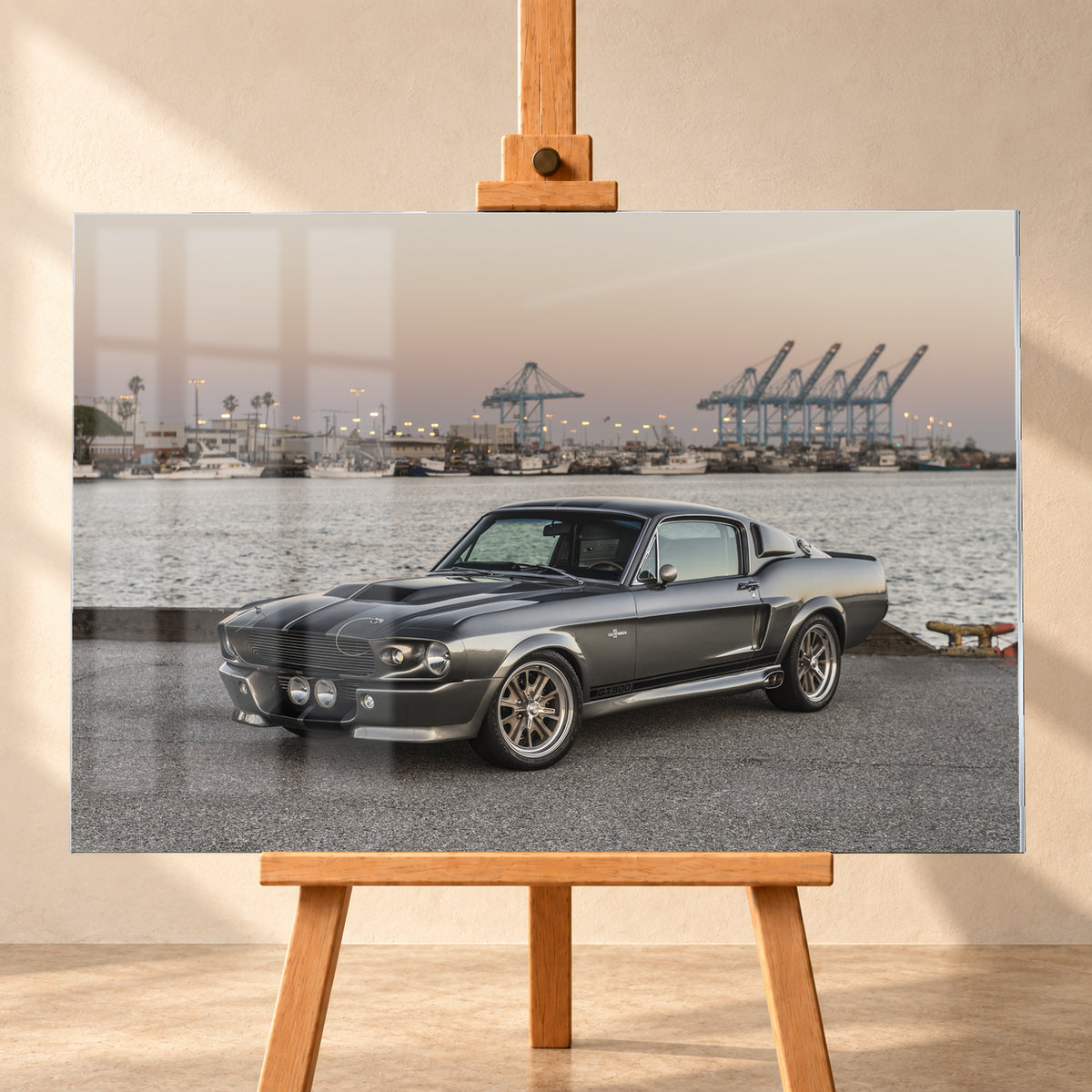 Dunkelgrauer Ford Mustang Shelby GT500 mit kontrastierenden Längsstreifen in Front-Dreiviertelansicht auf einer Hafenfläche; dahinter Wasser, Boote, Containerkräne, leuchtende Hafenlampen und ein pastellfarbener Abendhimmel; Visualisierung als Acrylglas-Wandbild, frontal auf einer Holzstaffelei.