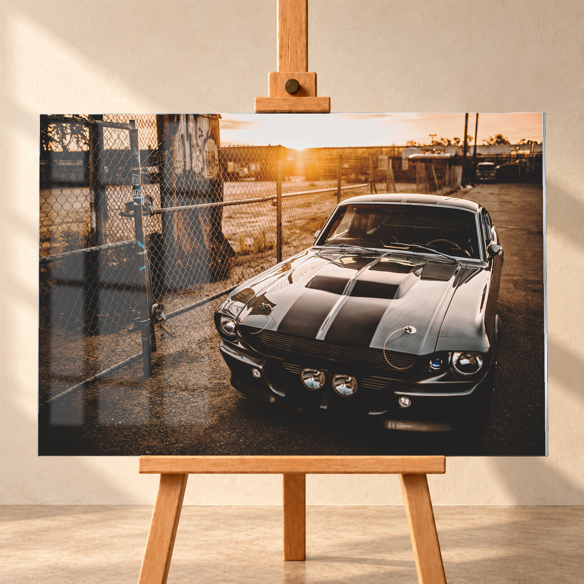 Silbergraues klassisches Fastback mit schwarzen Längsstreifen, schräg von vorn fotografiert, vor Maschendrahtzaun und Tor auf einem Industriegelände bei tief stehender, warm leuchtender Sonne; Visualisierung als Acrylglas-Wandbild, frontal auf einer Holzstaffelei.