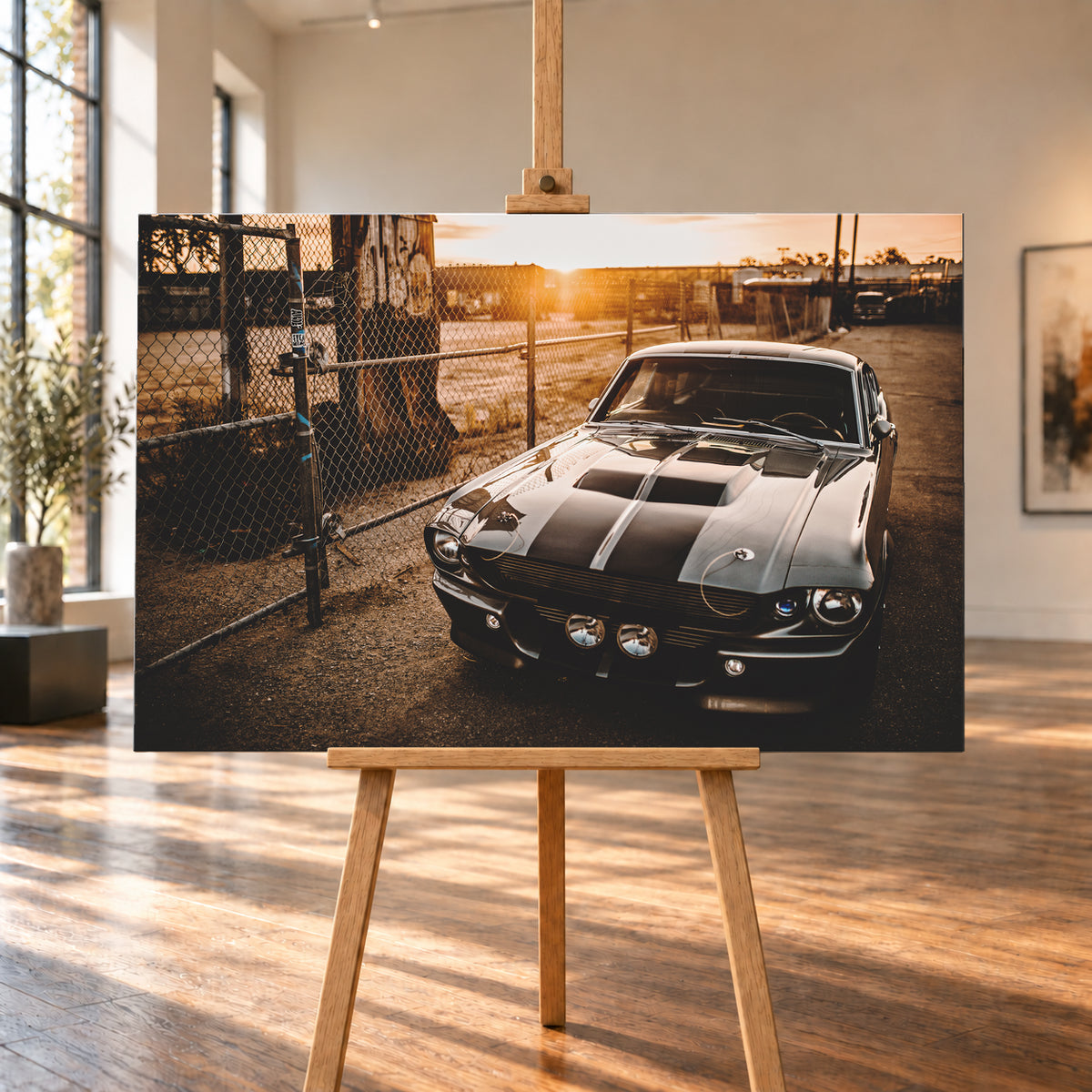 Silbergraues klassisches Fastback mit schwarzen Längsstreifen, schräg von vorn fotografiert, vor Maschendrahtzaun und Tor auf einem Industriegelände bei tief stehender, warm leuchtender Sonne; Visualisierung als Leinwand-Wandbild, auf einer Holzstaffelei in einem hellen Galerieraum.