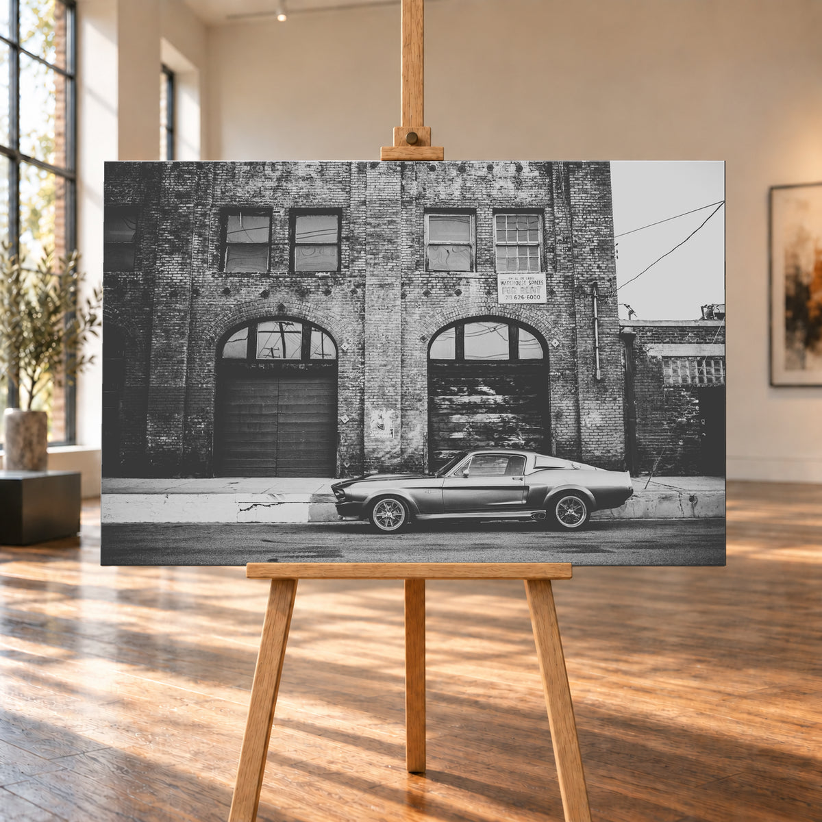Schwarz-Weiß-Aufnahme von Eleanor in vollständiger Seitenansicht vor einer verwitterten Backsteinfassade mit großen Toren und Fenstern an einer leeren Straße; Visualisierung als Leinwand-Wandbild, auf einer Holzstaffelei in einem hellen Galerieraum.