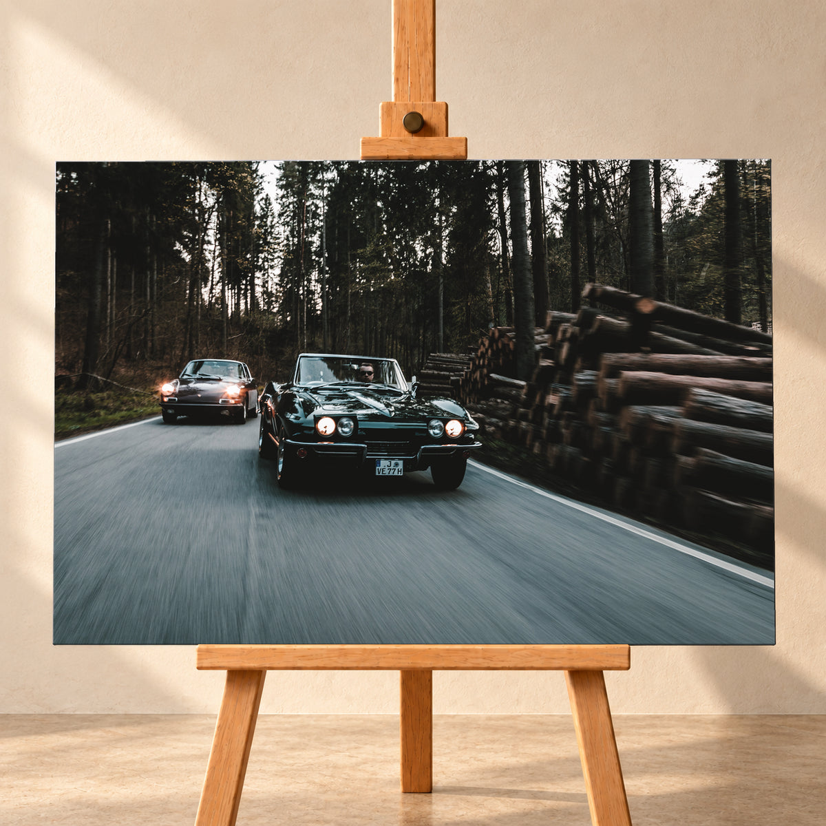 Eine dunkelgrüne 1967 Chevrolet Corvette C2 fährt frontal im Vordergrund einer Waldstraße; links dahinter ist ein dunkelroter 1967 Porsche 911 S Softwindow zu sehen. Die Scheinwerfer leuchten, Fahrer sind sichtbar. Hohe Bäume, gestapelte Baumstämme und Bewegungsunschärfe prägen die Szene; Visualisierung als Leinwand-Wandbild, frontal auf einer Holzstaffelei.
