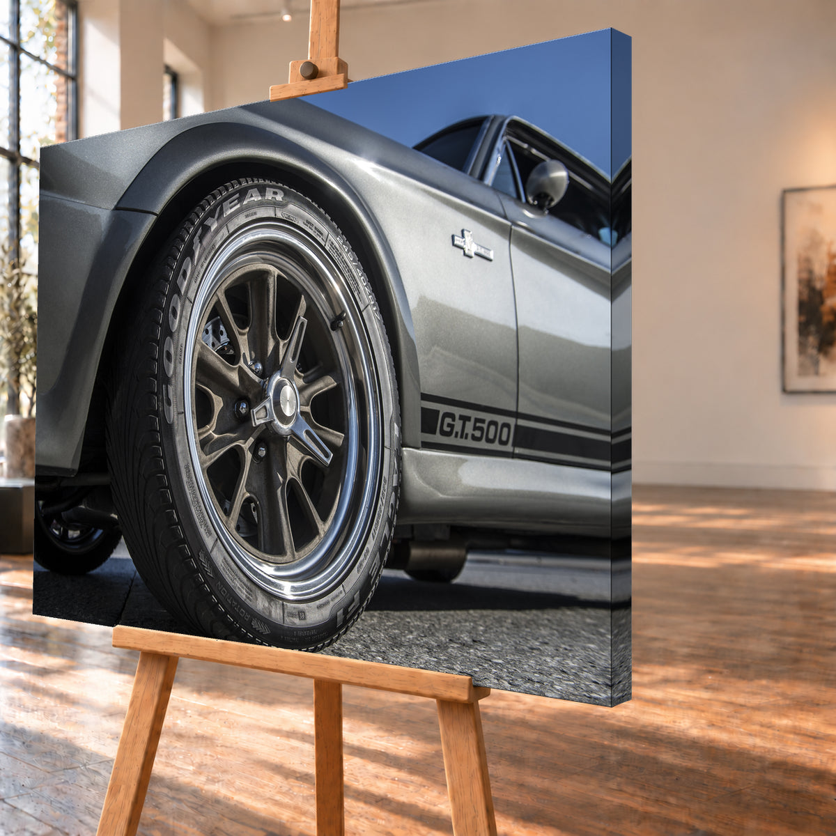 Nahaufnahme des Vorderrads eines silbergrauen Ford Mustang Shelby GT500 aus tiefer Perspektive. Dunkle Speichenfelge und Reifen dominieren das Bild; dahinter sind der Seitenstreifen mit „G.T.500“, Tür, Fenster, Außenspiegel und blauer Himmel zu sehen; Visualisierung als Leinwand-Wandbild, schräg von der Seite auf einer Holzstaffelei.