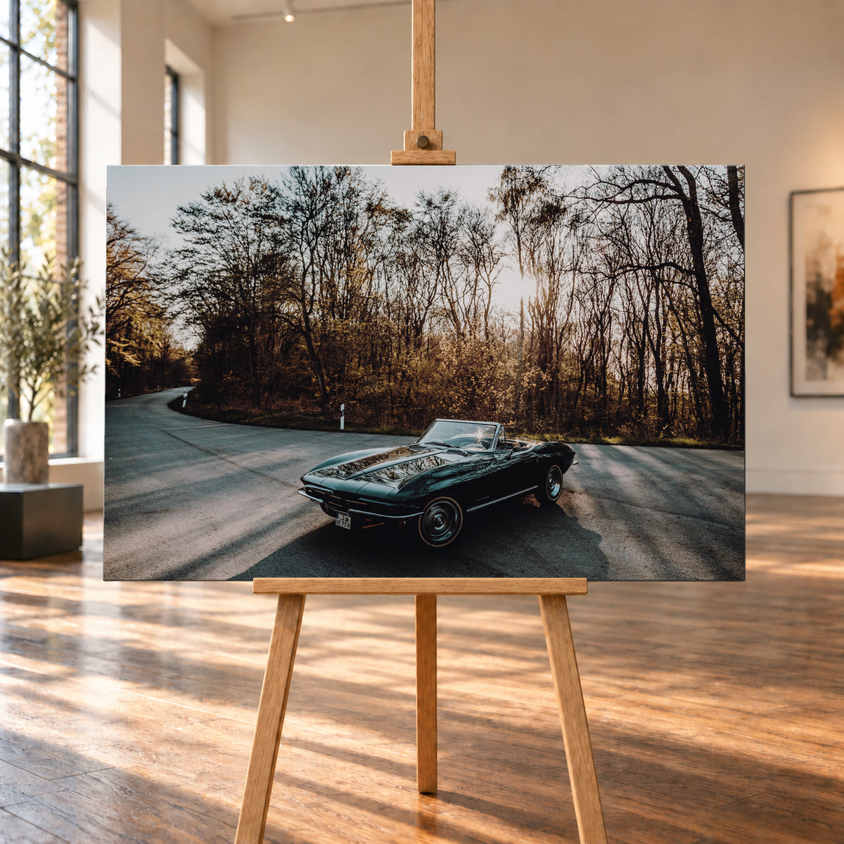Eine dunkelgrüne 1967 Chevrolet Corvette C2 als offenes Cabriolet in Front-Dreiviertelansicht auf einer kurvigen Waldstraße. Tief stehende Sonne scheint durch kahle Bäume; Gegenlicht und Astspiegelungen liegen auf der Karosserie; Visualisierung als Leinwand-Wandbild, auf einer Holzstaffelei in einem hellen Galerieraum.