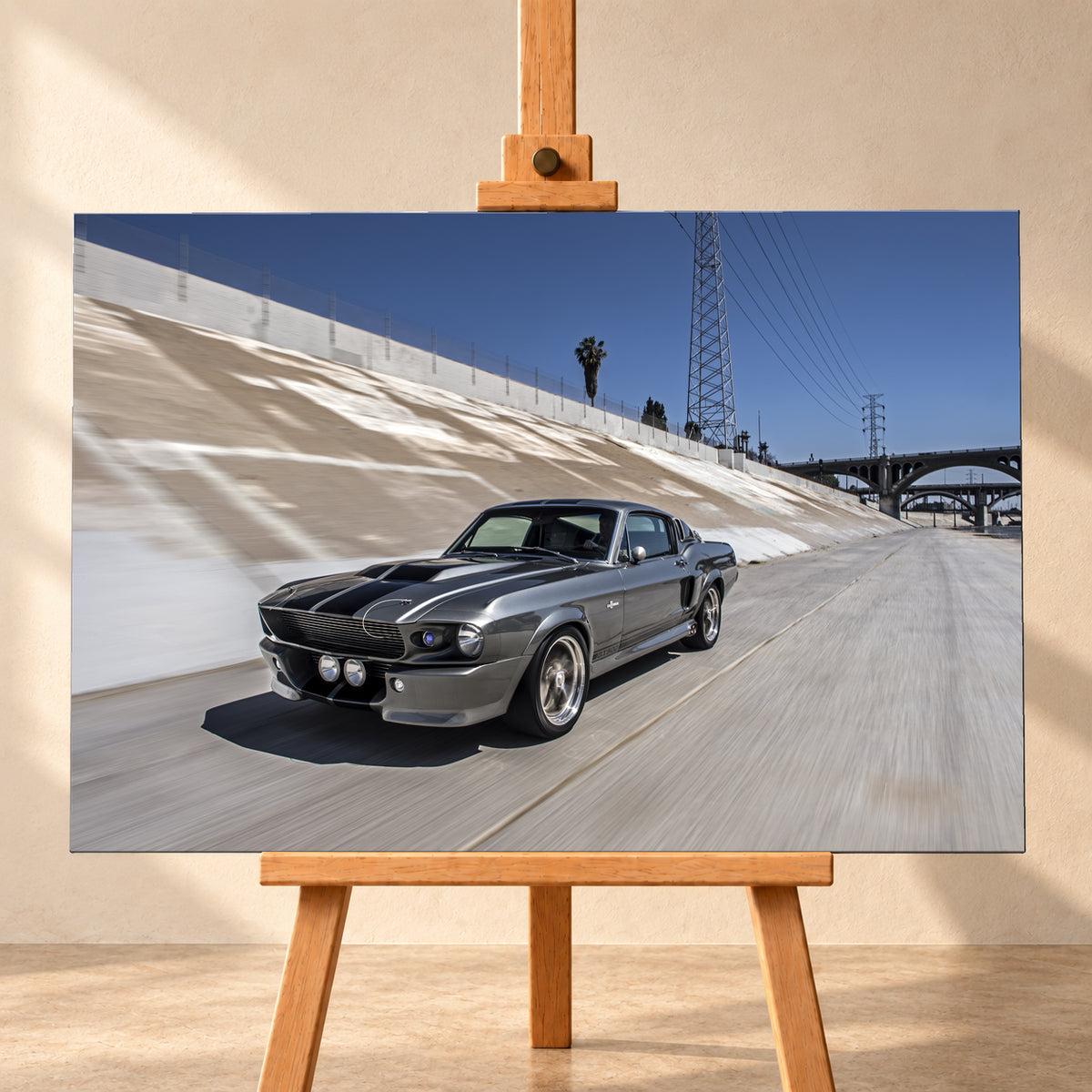 Metallisch grauer Ford Mustang Shelby GT500 mit schwarzen Doppelstreifen in niedriger Front-Dreiviertelansicht in einem hellen Betonkanal. Fahrbahn und Kanalwand sind bewegungsunscharf; Strommasten, Leitungen, Palmen und Bogenbrücken stehen unter blauem Himmel; Visualisierung als Leinwand-Wandbild, frontal auf einer Holzstaffelei.