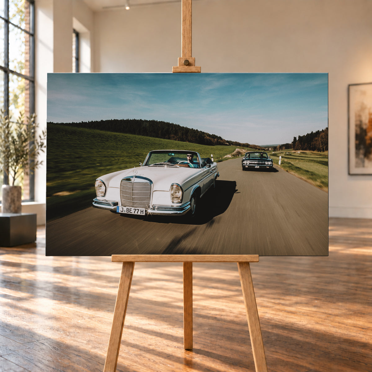 Frontale Fahraufnahme eines weißen 1965 Mercedes 300 SE W112 Cabriolets im Vordergrund und eines dunklen 1964 Lincoln Continental Cabriolets dahinter auf einer Landstraße. Grüne Felder, Waldlinien, blauer Himmel und Bewegungsunschärfe umgeben die beiden offenen Fahrzeuge im Sonnenlicht; Visualisierung als Leinwand-Wandbild, auf einer Holzstaffelei in einem hellen Galerieraum.