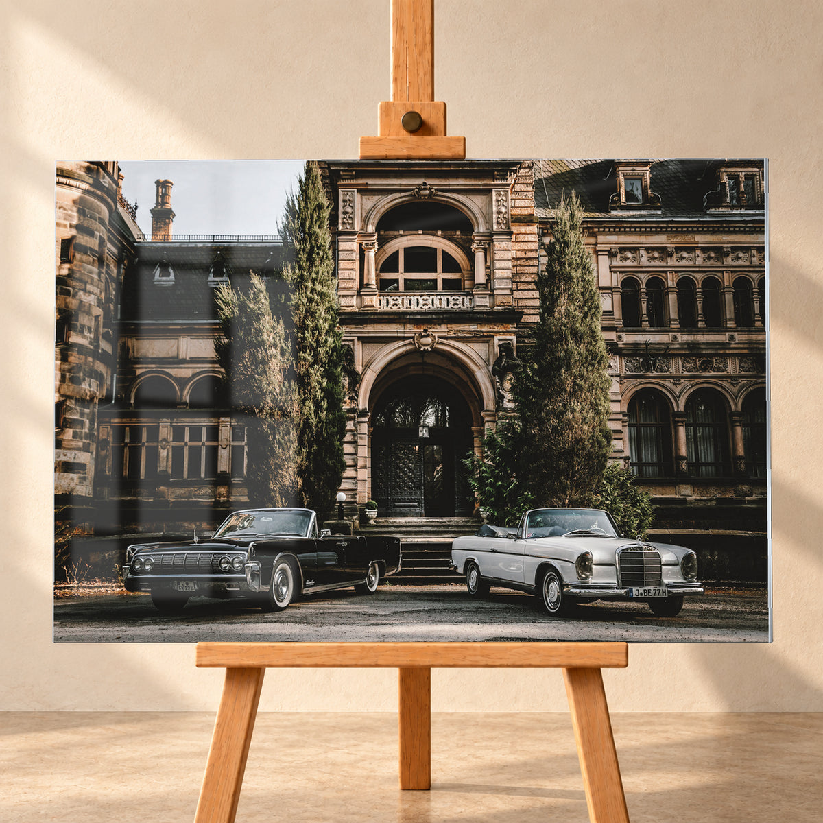Dunkler 1964 Lincoln Continental links und heller 1965 Mercedes 300 SE W112 rechts, beide als offene Cabriolets in schräger Frontansicht vor einer ornamentierten historischen Steinfassade mit Rundbögen, hohen Fenstern und schlanken Bäumen; Visualisierung als Acrylglas-Wandbild, frontal auf einer Holzstaffelei.