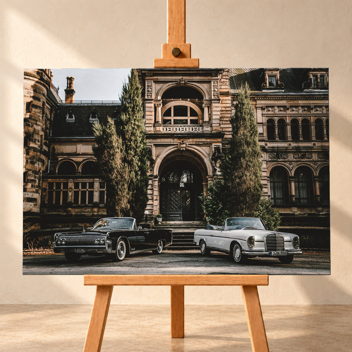 Dunkler 1964 Lincoln Continental links und heller 1965 Mercedes 300 SE W112 rechts, beide als offene Cabriolets in schräger Frontansicht vor einer ornamentierten historischen Steinfassade mit Rundbögen, hohen Fenstern und schlanken Bäumen; Visualisierung als Leinwand-Wandbild, frontal auf einer Holzstaffelei.