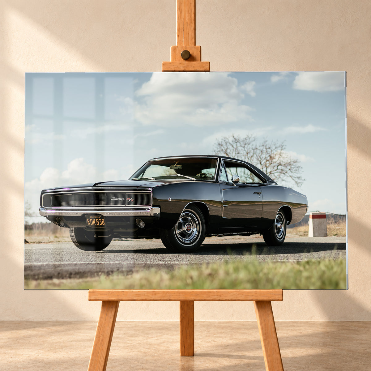 Dunkler Dodge Charger R/T in tiefer Dreiviertelansicht von vorn auf einer Straße unter hellem, wolkigem Himmel; im Hintergrund kahle Bäume und trockenes Gras; Visualisierung als Acrylglas-Wandbild, frontal auf einer Holzstaffelei.