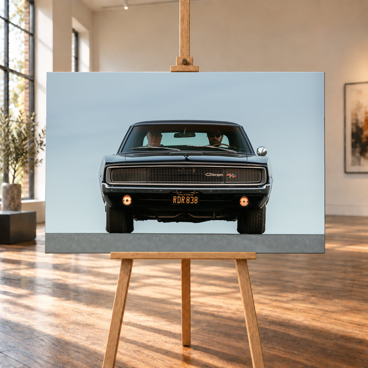Sehr dunkler Dodge Charger R/T in tiefer, zentraler Frontalansicht vor blassblauem Himmel; zwei Personen sitzen hinter der Windschutzscheibe, zwei Zusatzleuchten glimmen warm; Visualisierung als Leinwand-Wandbild, auf einer Holzstaffelei in einem hellen Galerieraum.