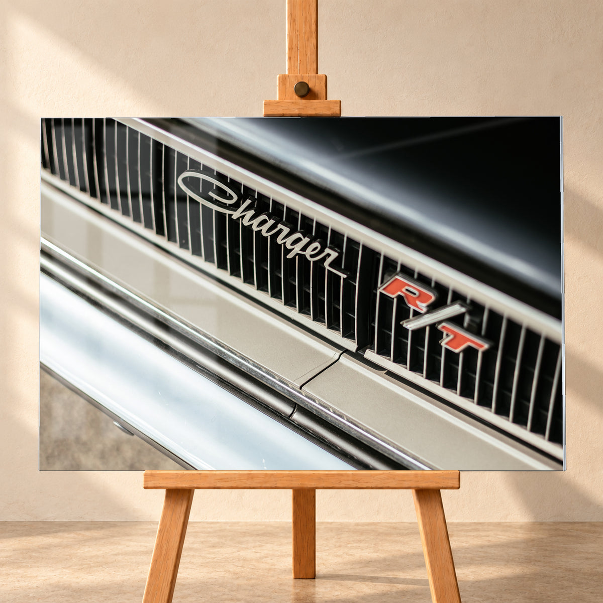 Diagonale Nahaufnahme eines hell lackierten Charger R/T mit schwarzem Lamellengrill, verchromtem Charger-Schriftzug und rot-weißem R/T-Emblem; Chromleisten reflektieren helles Licht; Visualisierung als Acrylglas-Wandbild, frontal auf einer Holzstaffelei.
