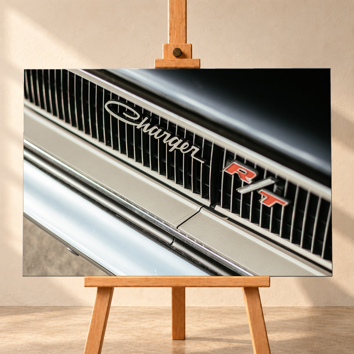 Diagonale Nahaufnahme eines hell lackierten Charger R/T mit schwarzem Lamellengrill, verchromtem Charger-Schriftzug und rot-weißem R/T-Emblem; Chromleisten reflektieren helles Licht; Visualisierung als Leinwand-Wandbild, frontal auf einer Holzstaffelei.
