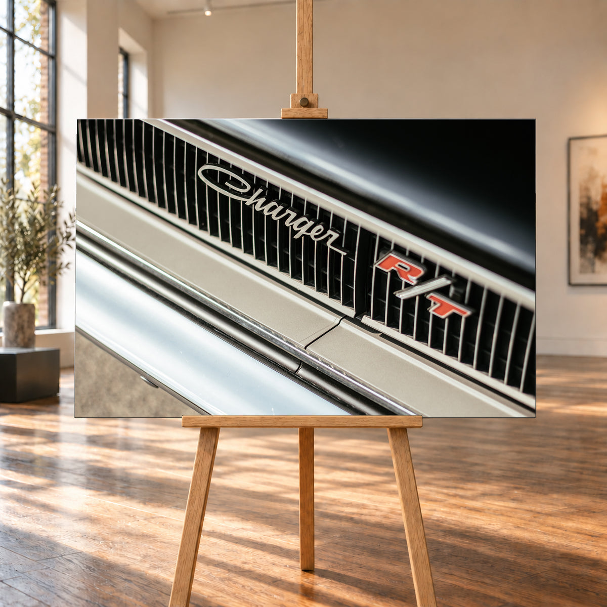Diagonale Nahaufnahme eines hell lackierten Charger R/T mit schwarzem Lamellengrill, verchromtem Charger-Schriftzug und rot-weißem R/T-Emblem; Chromleisten reflektieren helles Licht; Visualisierung als Leinwand-Wandbild, auf einer Holzstaffelei in einem hellen Galerieraum.