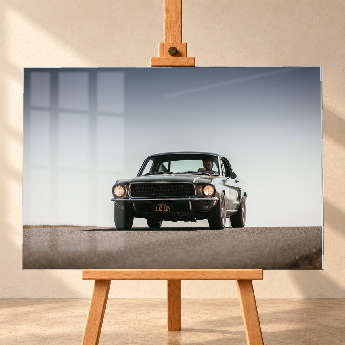 Dunkelgrüner klassischer Ford Mustang in tiefer Frontansicht auf Asphalt, mit eingeschalteten Rundscheinwerfern, sichtbarem Fahrer und weitem hellgrauem Himmel; Visualisierung als Acrylglas-Wandbild, frontal auf einer Holzstaffelei.