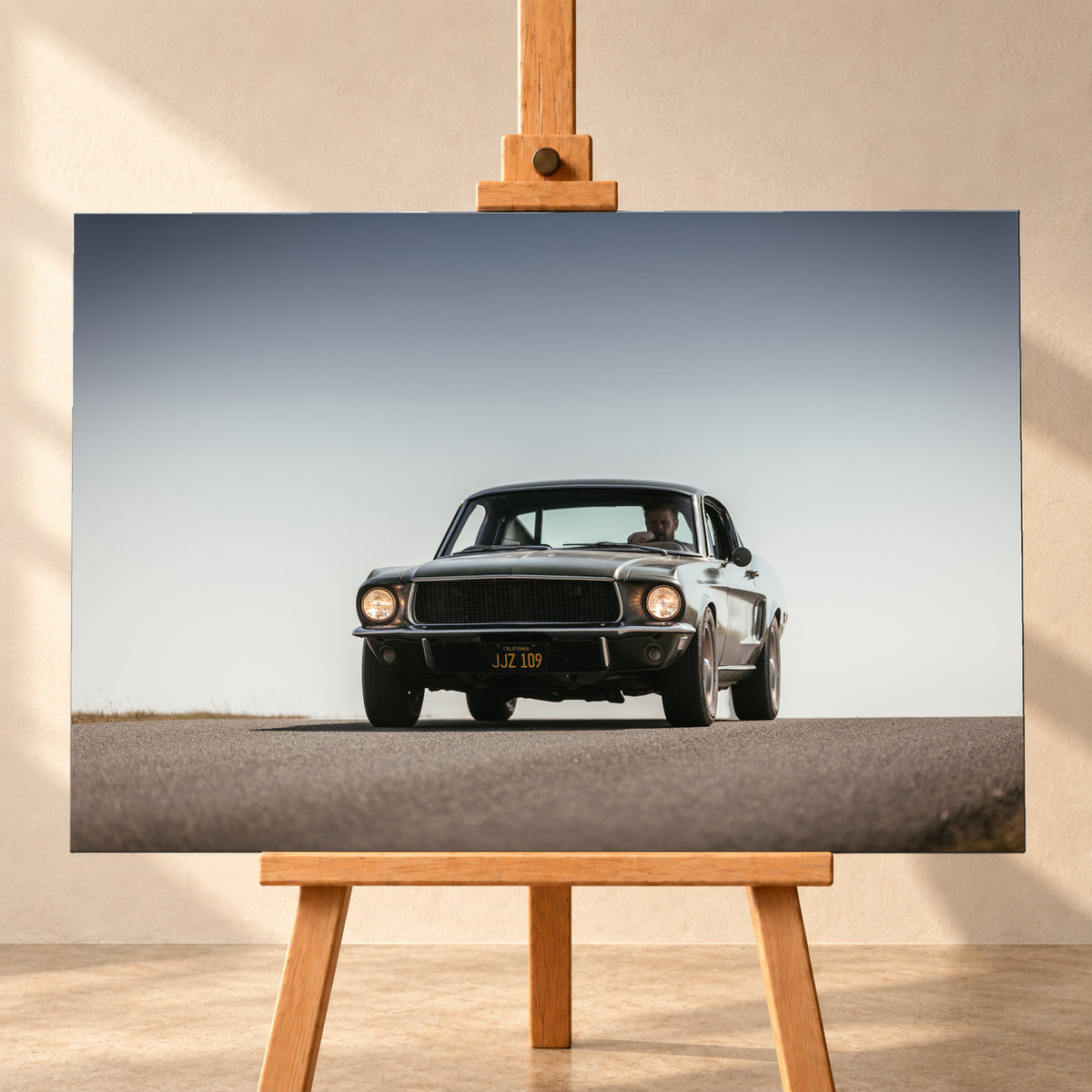 Dunkelgrüner klassischer Ford Mustang in tiefer Frontansicht auf Asphalt, mit eingeschalteten Rundscheinwerfern, sichtbarem Fahrer und weitem hellgrauem Himmel; Visualisierung als Leinwand-Wandbild, frontal auf einer Holzstaffelei.
