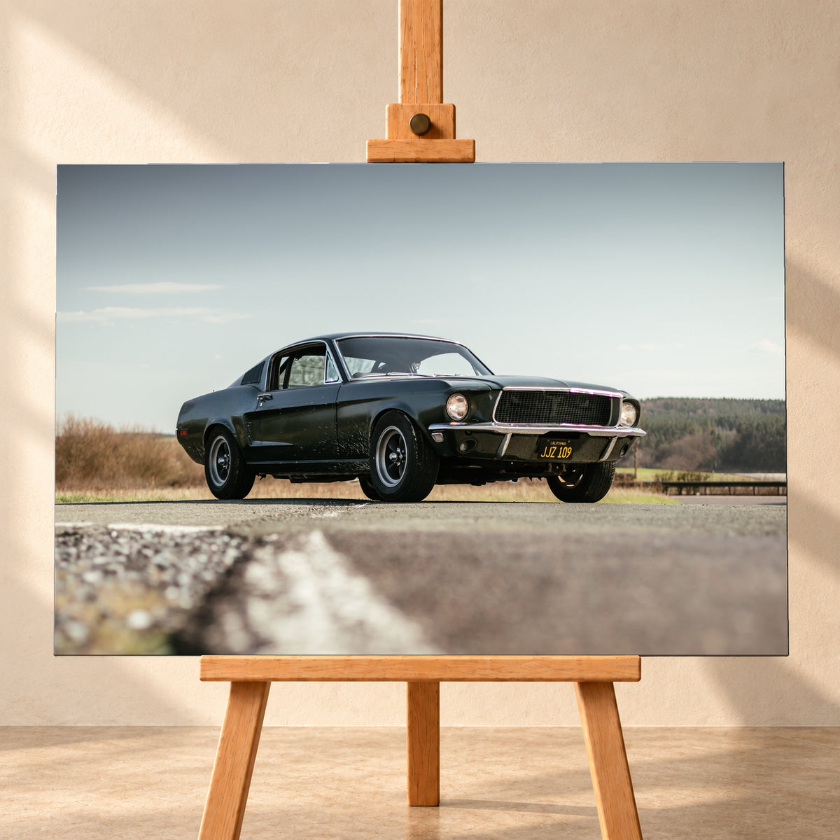 Dunkelgrüner Ford Mustang in niedriger Dreiviertelansicht von vorn auf Asphalt; dahinter offene Felder, ein bewaldeter Horizont und heller Himmel bei Tageslicht; Visualisierung als Leinwand-Wandbild, frontal auf einer Holzstaffelei.