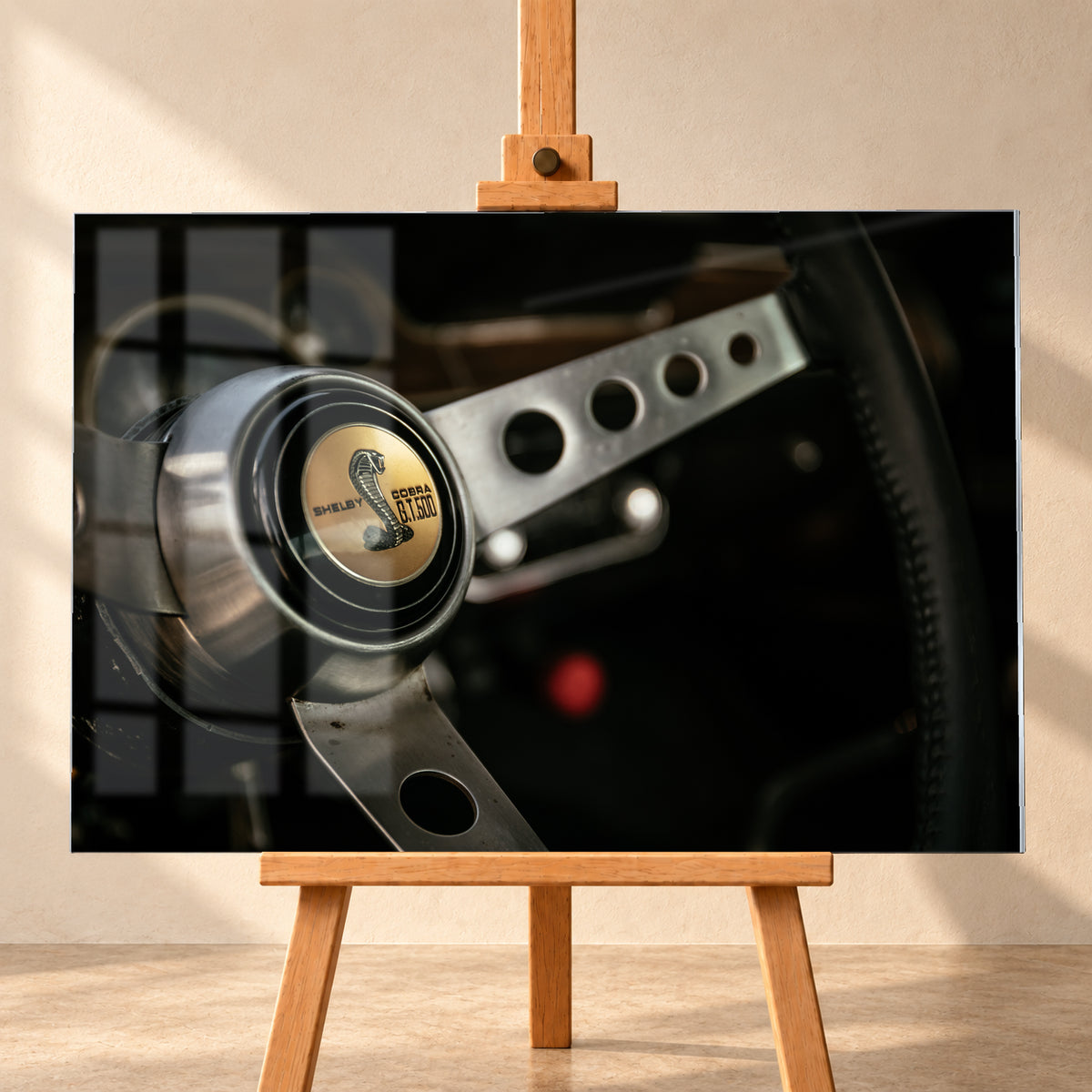 Nahaufnahme eines dreispeichigen Lenkrads mit gelochten silberfarbenen Speichen, schwarzem Kranz und goldenem Shelby-Cobra-GT500-Emblem in einem dunklen, warm beleuchteten Cockpit; Visualisierung als Acrylglas-Wandbild, frontal auf einer Holzstaffelei.