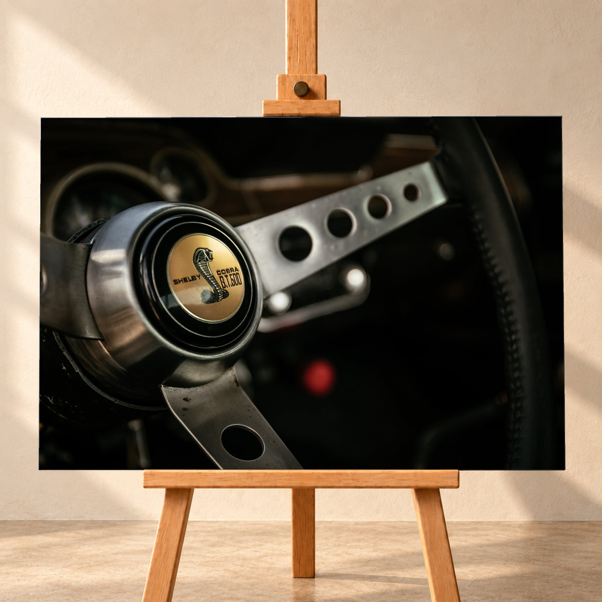 Nahaufnahme eines dreispeichigen Lenkrads mit gelochten silberfarbenen Speichen, schwarzem Kranz und goldenem Shelby-Cobra-GT500-Emblem in einem dunklen, warm beleuchteten Cockpit; Visualisierung als Leinwand-Wandbild, frontal auf einer Holzstaffelei.