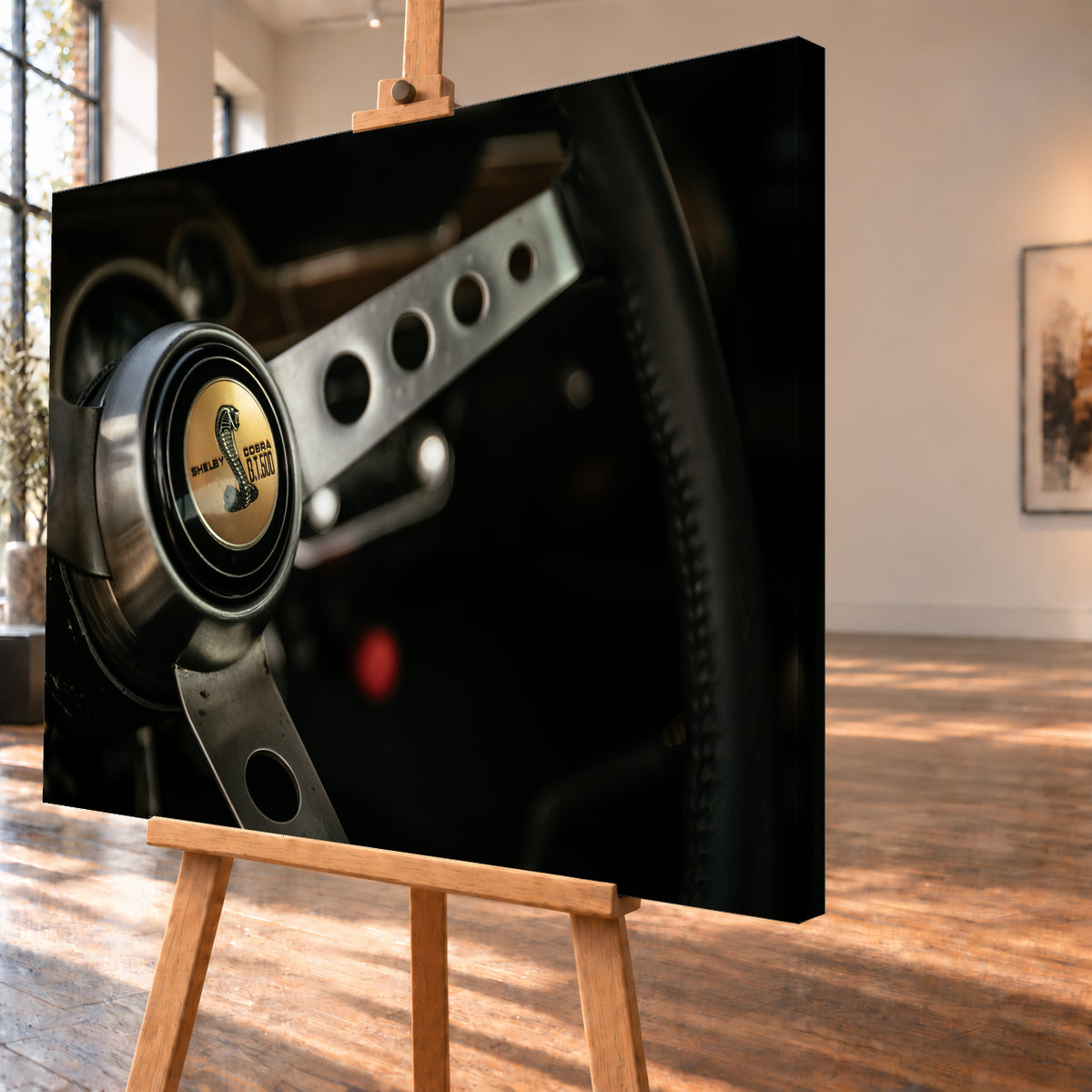 Nahaufnahme eines dreispeichigen Lenkrads mit gelochten silberfarbenen Speichen, schwarzem Kranz und goldenem Shelby-Cobra-GT500-Emblem in einem dunklen, warm beleuchteten Cockpit; Visualisierung als Leinwand-Wandbild, schräg von der Seite auf einer Holzstaffelei.