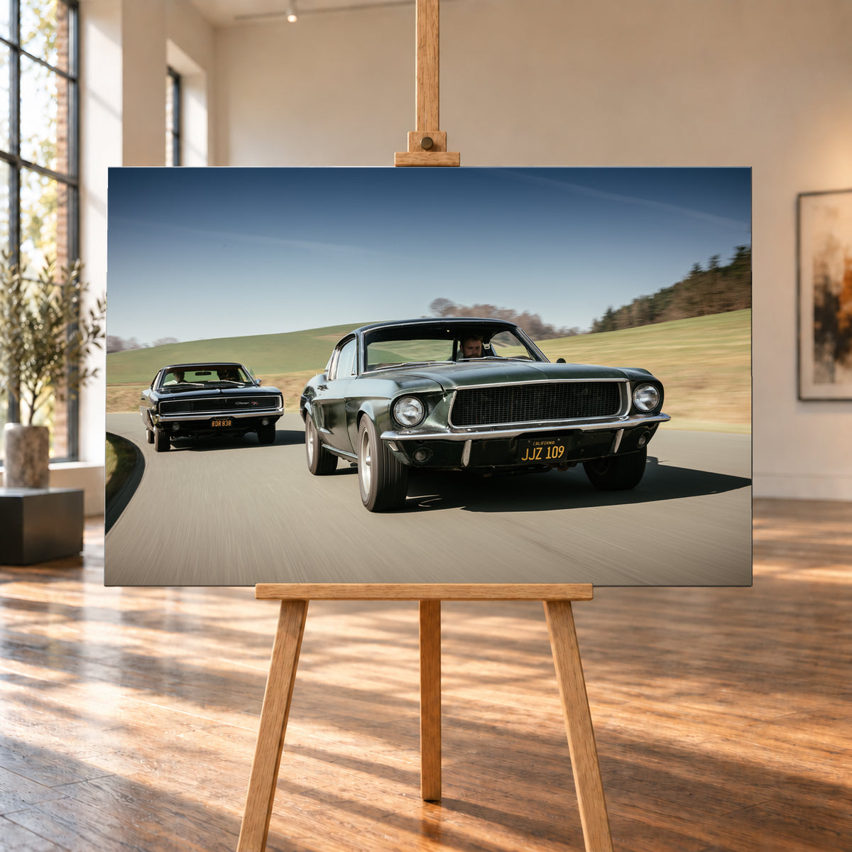 Dynamische Fahraufnahme zweier klassischer Coupés auf einer kurvigen Landstraße. Im Vordergrund fährt ein dunkelgrünes Coupé in Front-Dreiviertelansicht; links dahinter folgt der dunkle Bullitt Charger. Grüne Hügel, Bäume und blauer Himmel rahmen die helle Szene, während verwischte Straße und Umgebung Bewegung vermitteln; Visualisierung als Leinwand-Wandbild, auf einer Holzstaffelei in einem hellen Galerieraum.