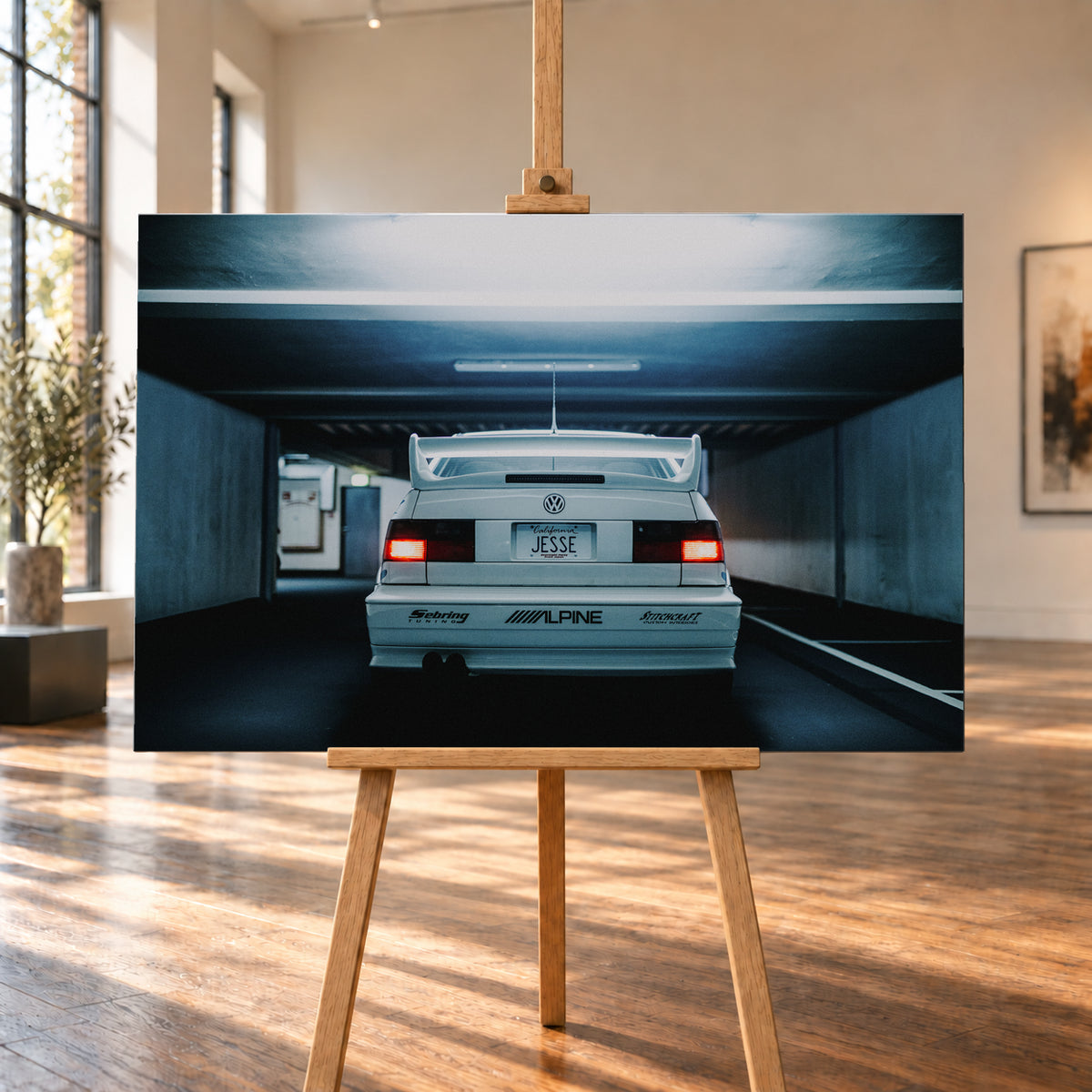 Weißer Volkswagen Jetta in symmetrischer Heckansicht in einer dunklen Betongarage, mit großem Heckflügel, roten Rücklichtern, sichtbaren Aufklebern und kühlem blau-grauem Licht; Visualisierung als Leinwand-Wandbild, auf einer Holzstaffelei in einem hellen Galerieraum.
