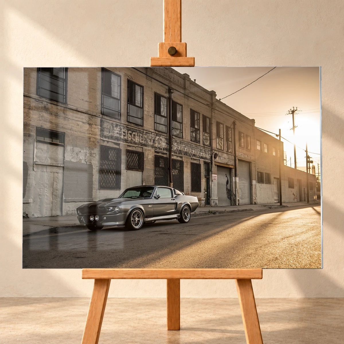 Frontale Visualisierung des Acrylglas-Wandbilds auf einer Holzstaffelei vor heller Wand. Das Motiv zeigt den silbergrauen Mustang „Eleanor“ vor verwitterten Industriegebäuden bei tief stehender Sonne. Dargestellte Fensterreflexionen überlagern links das Motiv.