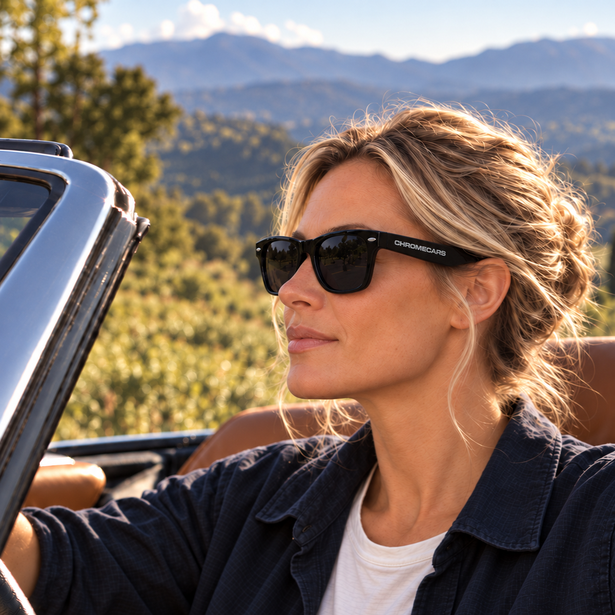 Frau mit schwarzer Sonnenbrille und sichtbarem CHROMECARS-Schriftzug in einem offenen Auto; Bäume und Berge liegen im Hintergrund.
