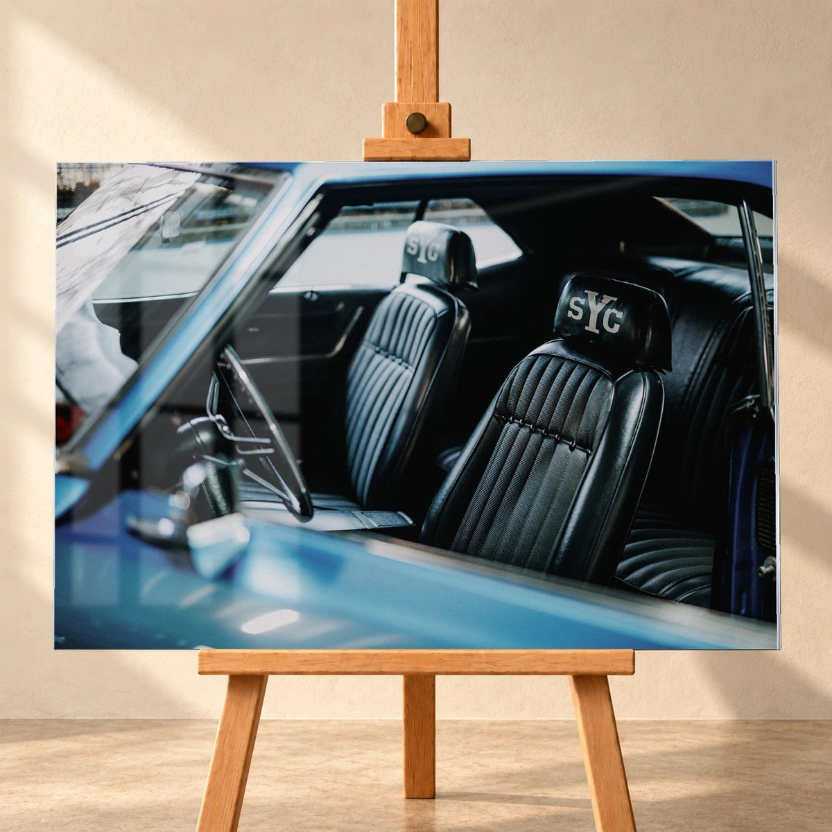 Frontale Visualisierung eines Acrylglasbildes im Querformat auf einer Holzstaffelei vor heller Wand. Zu sehen ist der Innenraum des blauen Chevrolet Camaro Yenko mit schwarzen Vordersitzen, weißen „sYc“-Schriftzügen auf den Kopfstützen und dem Lenkrad links.