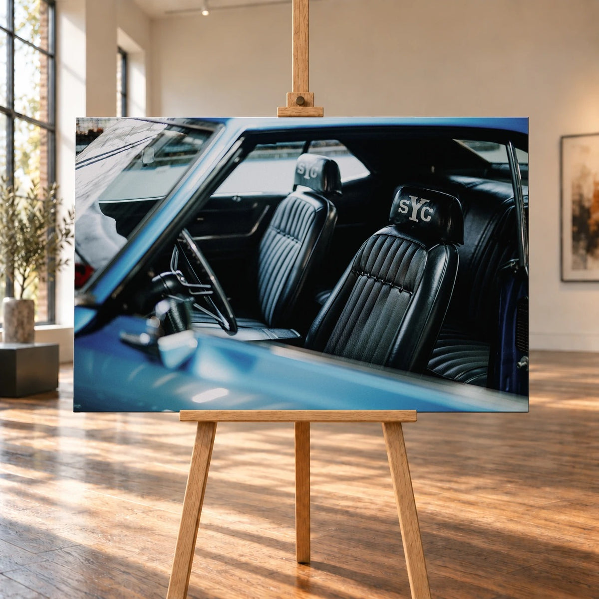 Raumvisualisierung mit einem querformatigen Leinwandbild auf einer Holzstaffelei in einem hellen Raum mit großen Fenstern und Holzboden. Abgebildet ist der Innenraum des blauen Chevrolet Camaro Yenko mit schwarzen Sitzen und „sYc“-Schriftzügen auf den Kopfstützen.