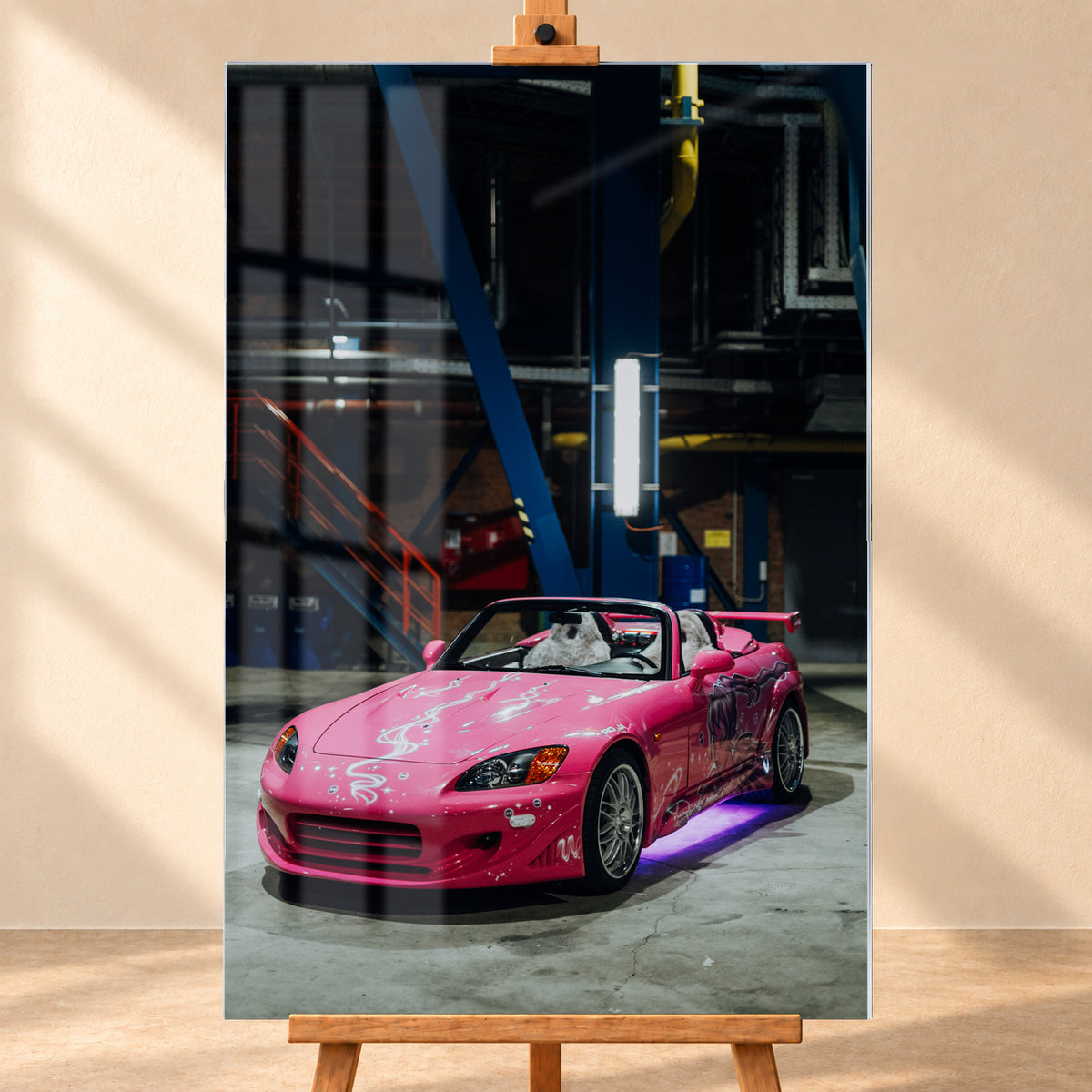 Frontale Produktvisualisierung eines Acrylglasbilds im Hochformat auf einer Holzstaffelei vor heller Wand. Fensterreflexionen überlagern das Motiv des pinken Honda S2000 aus „2 Fast 2 Furious“, der schräg von vorn in einer dunklen Industriehalle zu sehen ist.
