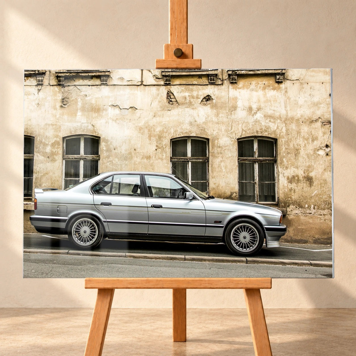 Frontale Visualisierung eines Acrylglas-Wandbilds auf einer Holzstaffelei vor heller Wand. Lichtreflexe liegen über dem Motiv eines silbernen BMW Alpina B10, der seitlich vor einer verwitterten Fassade mit dunklen Fenstern gezeigt wird.