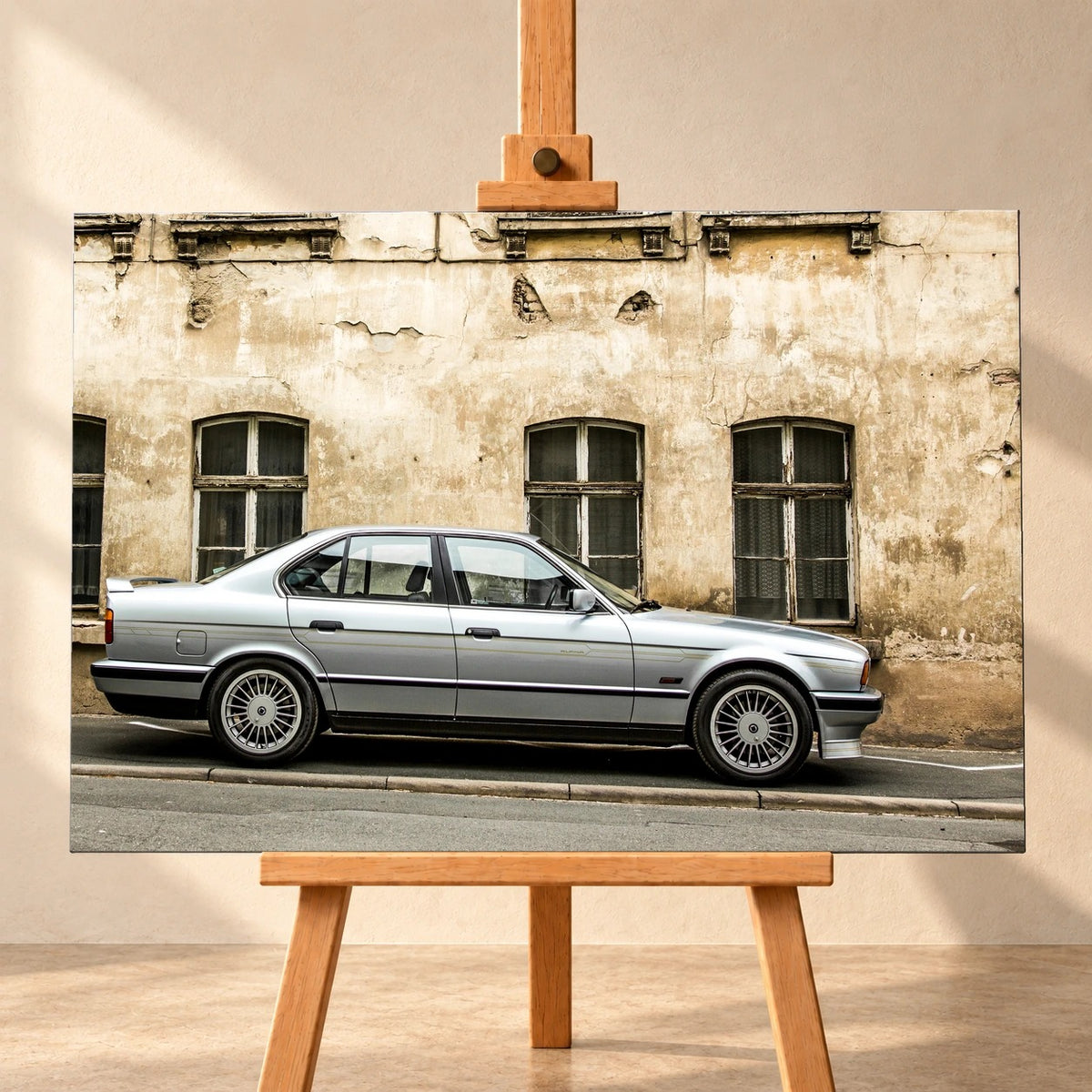 Visualisierung eines Leinwandbilds im Querformat auf einer Holzstaffelei vor heller Wand. Das Motiv zeigt einen silbernen BMW Alpina B10 in Seitenansicht vor einer verwitterten Fassade mit hohen, dunklen Fenstern.
