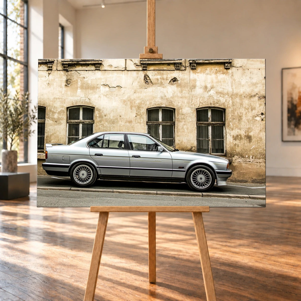 In dieser Raumvisualisierung steht das querformatige Leinwandbild auf einer Holzstaffelei in einem hellen Raum mit großen Fenstern und Holzboden. Auf dem Bild ist ein silberner BMW Alpina B10 im Profil vor einer verwitterten Hausfassade zu sehen.